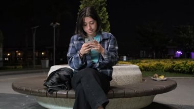 Parkta tek başına oturup telefonuna bakan, dokunmatik ekranda parmaklarını hızla kullanan genç bir kızın akşam vakti parkta tek başına vakit geçirdiği görüntüleri.