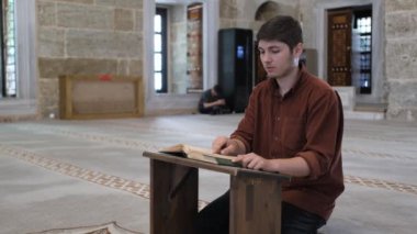 Diz çöküp Kuran okuyan genç adam caminin içindeki ahşap tutacağa farklı açılardan bakıyor camide İslam 'ın kutsal kitabını okuyor.