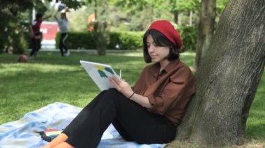 Parkta yeşil ağaca yaslanmış güzel bir kadın, beyaz tuvalde fırçayla resim yapıyor, açık havada renkli boyalarla resim yapıyor.