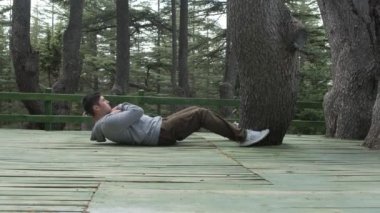 Yeşil orman bankında spor yapan genç bir adamın görüntüsü, ormanda egzersiz yaparak güçleniyor, çam ağaçları arasında mekik çeken bir adam.
