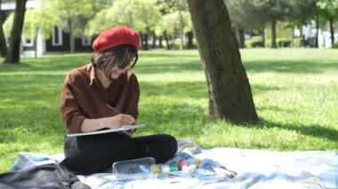 Zengin ağaçların arasında oturan ve resim yapan kadın sanatçı, açık havada fırçayla çizim yapan, kırmızı şapkalı sanatçı çimenlerde oturuyor ve doğayı resmediyor.