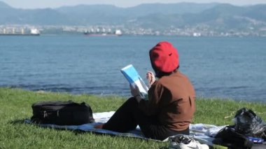 Deniz kenarında oturup resim yapan, dışarıda resim yapan, deniz manzarasına karşı yeşil çimlere resim çizen güzel bir kız resmi.