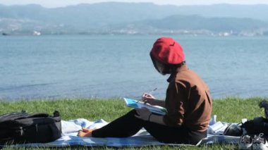 Deniz manzarası ve resim karşısında oturan genç kadın ressam, deniz kenarındaki çimenlerde oturan ve tuvale resim çizen kadın.