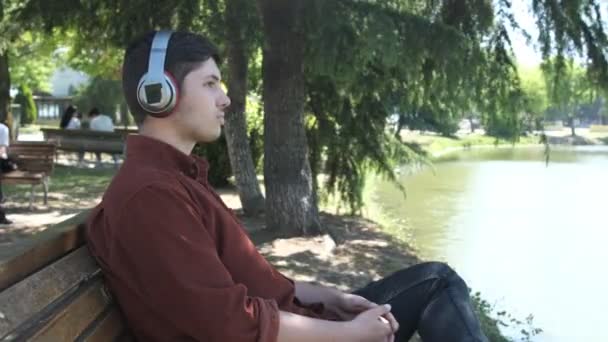 緑の木々の下に座ってヘッドフォンで音楽を聴いている若い男は 湖のそばのバンドに座って見て 緑の風景の中で楽しい時間 — ストック動画