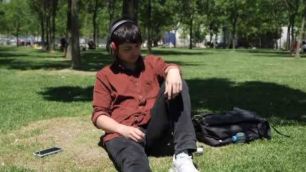 晴れた日に音楽を聴くことによって木にもたれ リラックスした人 芝生の上で歌を聴く少年のイメージ 一般的なヘッドセットで音楽を聴くティーン — ストック動画