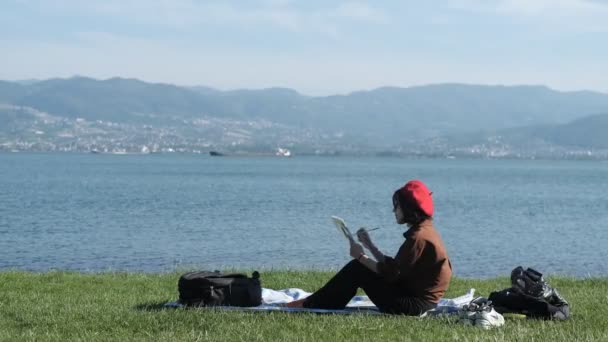 美丽的女孩坐在海边看风景 在外面作画 在碧绿的草地上对着海景作画 — 图库视频影像