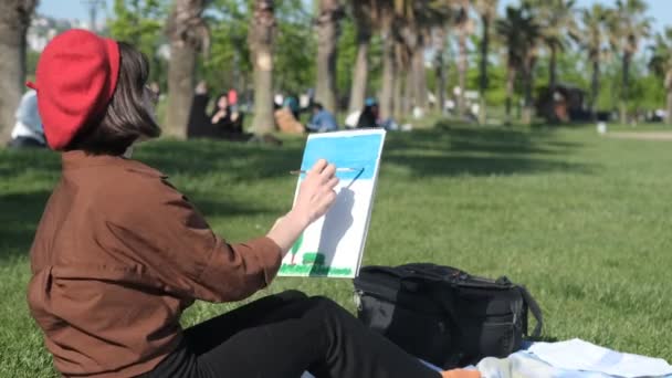 緑の公園の地面に座っている若い女性の絵のイメージ 芝生の上に座っている赤い帽子のアーティスト キャンバス上の絵 — ストック動画