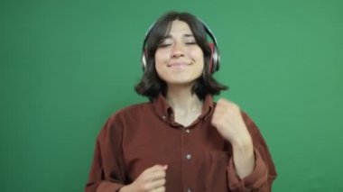 Müzik dinleyen, kırmızı kulaklıklarla eğlenen, müzik eşliğinde dans eden güzel bir kız, yeşil perdenin önünde genç bir kadının yüz ifadesi.