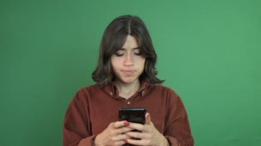 Image of girl texting nervously on the phone, young girl looking at the phone with an angry facial expression, facial expression of young woman in front of green curtain