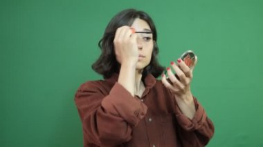 Young girl doing makeup with a mirror in her hand, image of a girl using makeup to decorate, facial expression of young woman in front of green curtain