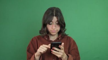 Image of young girl shocked at what she looks at while browsing the phone, image of surprised facial expression, facial expression of young woman in front of green curtain