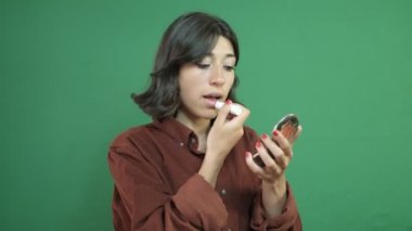 Young girl applying make-up in front of a green curtain, woman applying lipstick with a mirror, facial expression of young woman in front of green curtain