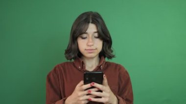 Elinde telefonla mesajlaşan, mesajlaşmadan sonra yüz ifadesini bekleyen genç bir kadının yeşil perdenin önündeki yüz ifadesi.