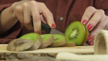 Tahta sunum tahtasında yeşil kivilerin dilimlenmesi, kadının kiviyi yakından kesmesi, tematik taze meyve hazırlama, ahşap sunum tabağında dilimleme işlemi.