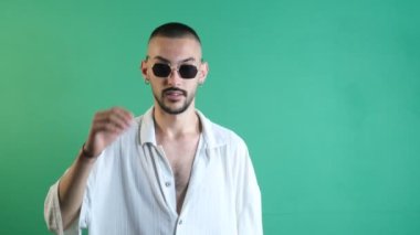 Gözlüklerini çıkaran bir adam, güneş gözlüğünü çıkaran havalı bir adam, yeşil ekranın önündeki modelin modaya uygun ve izole bir görüntüsü olması için beyaz gömlekli genç bir adam güneş gözlüğü takıyor.