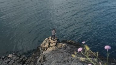 Deniz kıyısındaki kayalıklarda balık tutan bir adam resmi, siyah tişörtlü bir adam olta ipi kullanıyor, mavi deniz manzaralı bir balık.