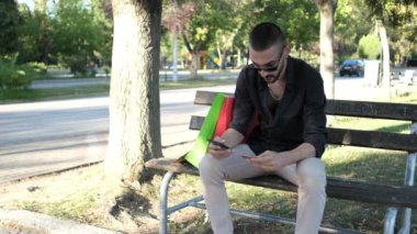 Online shopping with credit card, sitting on bench in park shopping on phone, online shopping from sitting place with red green shopping bags
