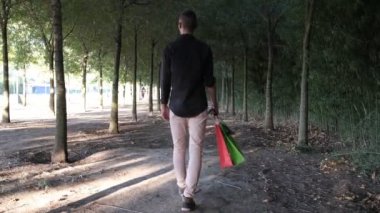 Man returning from shopping, young man returning from shopping with colorful bags in hand, walking on tree lined road after shopping