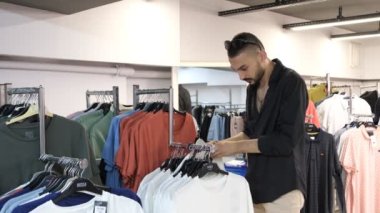 Man taking pictures while shopping, man trying on white t-shirt, man trying product in cabin to buy, reviewing and liking clothes hanging on hangers