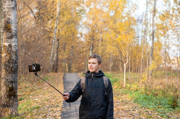 Хлопчик-підліток в чорній куртці і монопод в руці знімає відео на смартфоні. Хлопчик-блогер. Пройдіться через осінній парк. Крупним планом . — стокове фото