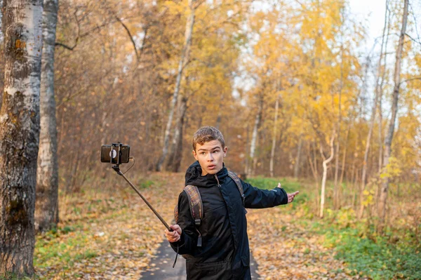 Хлопчик-підліток в чорній куртці і монопод в руці знімає відео на смартфоні. Хлопчик-блогер. Пройдіться через осінній парк. Крупним планом . — стокове фото