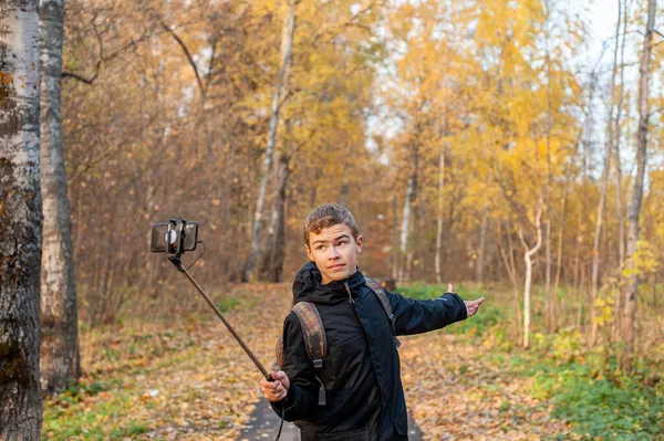 Een tiener in een zwart jasje en een monopod in zijn hand maakt video 's op een smartphone. Jongensblogger. Loop door het herfstpark. Close-up. — Stockfoto