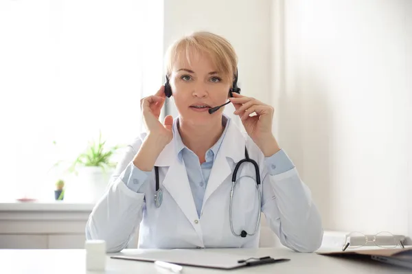 Mujer médico general lleva una bata blanca y auriculares hablando videoconferencia en el ordenador portátil utilizando la aplicación de consulta de videollamadas en línea. Asistencia médica remota para el paciente a distancia, telemedicina . — Foto de Stock