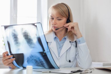 Beyaz önlüklü ve kulaklıklı ciddi bir doktor kadın akciğerlerinin röntgenini çekiyor. Terapist konsültasyon yapar ve teşhisi açıklar. Teletıp konsepti.