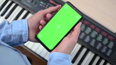 Elektronik piyano masasının üzerinde dikey yeşil ekranlı cep telefonu tutan bir gencin ya da gencin yakın çekimi. Müzik mesafesi öğrenme konsepti. Krom anahtar telefon ekranına odaklan