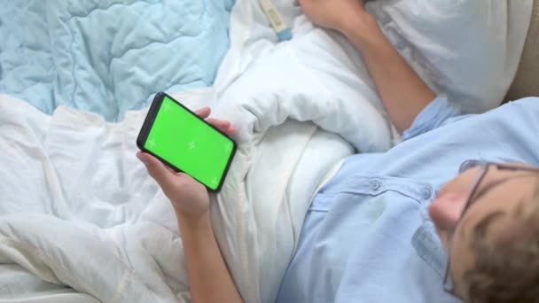 O adolescente se senta na cama sob as capas e segura um telefone celular com uma tela verde vertical e um termômetro eletrônico. O conceito de telemedicina. Concentre-se na tela do telefone — Vídeo de Stock