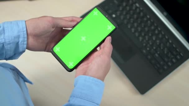 Close-up de um jovem ou adolescente mão segurando um telefone celular com uma tela verde vertical sobre uma mesa com um laptop. Concentre-se na tecla chroma da tela do telefone, — Vídeo de Stock