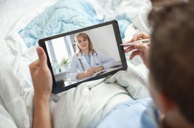 Teletıp ve online danışmanlık konsepti. Genç bir adam ya da genç bir adam elinde bir tablet ve elektronik termometreyle yatakta oturuyor. Genç adam bir doktorla görüntülü konuşma yapıyor..