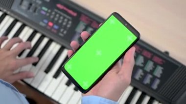 Elektronik piyano masasının üzerinde dikey yeşil ekranlı cep telefonu tutan bir gencin ya da gencin yakın çekimi. Müzik mesafesi öğrenme konsepti. Krom anahtar telefon ekranına odaklan