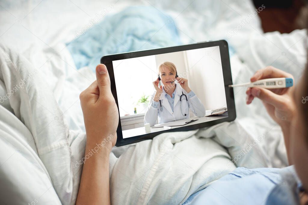 Telemedicine and online consultation concept. A young man or teenager sits in bed with an electronic thermometer and a tablet in his hands. The young man has video chat with a doctor.