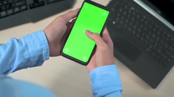 Detailní záběr mladého muže nebo mladistvých rukou držících mobilní telefon se svislou zelenou obrazovkou nad stolem s laptopem. Zaměření na tlačítko chroma obrazovky telefonu, — Stock video