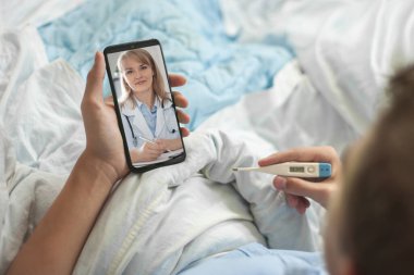 Teletıp ve online danışmanlık kavramı. Genç bir adam ya da ergen, elinde bir akıllı telefon ve elektronik termometreyle yatakta oturuyor. Genç bir adam doktorla görüntülü konuşma yapıyor..