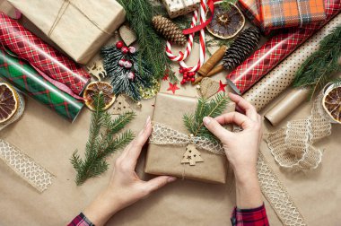 Kadınların elleri bir Noel hediyesi paketleyip süsler. Masa bej el işi kağıtlarla kaplı ve kırmızı ve yeşil renklerin dekorasyonu için malzemelerle dolu. Yukarıdan görüntüle