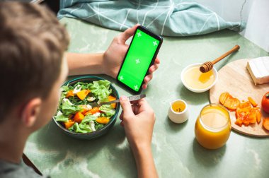 Kalori sayma, online beslenme alışverişi ve online eğitim kavramı. Genç bir çocuk sağlıklı bir vitamin salatasıyla kahvaltı yapar ve elinde yeşil renkli ekranlı bir telefon tutar..