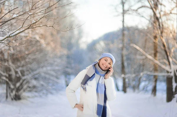 Beyaz önlüklü ve mavi şapkalı çekici, gülen sarışın bir kadın kışın karla kaplı bir parkta yürüyor ve telefonda konuşuyor. Aktif ve sağlıklı yaşam tarzı.