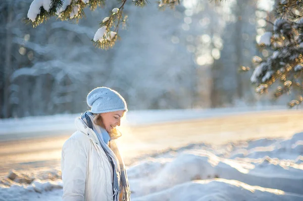  Beyaz önlüklü, mavi eşarplı ve şapkalı mutlu, gülen sarışın kadın, dondurucu güneşli bir günde ormanda yürüyor. Çam ağacının dallarından kar yağar Sağlıklı ve aktif yaşam tarzı Kış Yürüyüşü.
