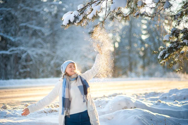 白いコートに青いスカーフと帽子で金髪の女性を笑って幸せな晴れた日に森の中を歩く。松の木の枝から雪が降る健康的で活発なライフスタイル冬の散歩. — ストック写真