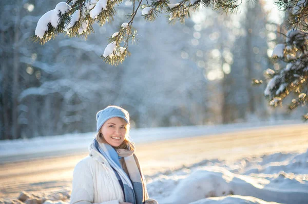 Happy laughing blonde woman in a white coat, a blue scarf and hat walks through the forest on frosty sunny day. Portrait of an attractive woman in winter clothes Healthy and active lifestyle — Stock Photo, Image