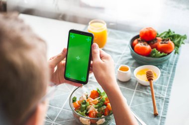 Kalori sayma, online beslenme alışverişi ve online eğitim kavramı. Genç bir çocuk sağlıklı bir vitamin salatasıyla kahvaltı yapar ve elinde yeşil renkli ekranlı bir telefon tutar..