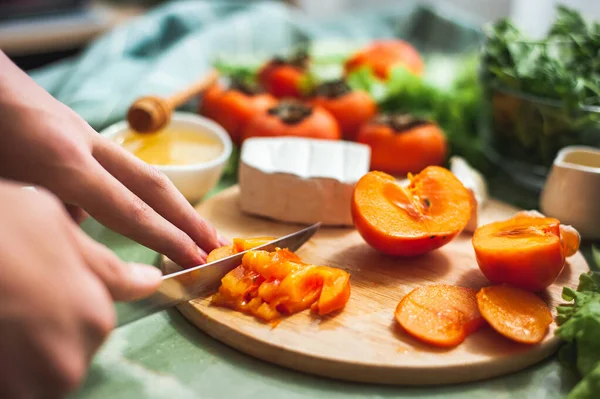 Ingredientes y proceso de hacer una ensalada de vitaminas de invierno con caquis, mandarinas y queso Cortes a mano femeninos o adolescentes caquis en una tabla redonda de madera Preparación para el almuerzo vegano. Enfoque selectivo — Foto de Stock