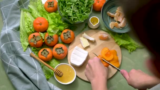 Příprava veganského salátu z persimonů, mandarinek a salátu. Pohled shora. Pohyb kamery. Lidské ruce plátky ovoce pro zdravou snídani nebo oběd. — Stock video