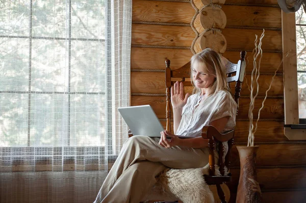 Uma mulher loira em roupas de casa bege senta-se em uma cadeira de balanço com um laptop em suas mãos em um quarto acolhedor com grandes janelas e paredes de madeira. Freelancing e ensino à distância. — Fotografia de Stock