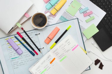 Öğrencilerin çalışma alanı. Masanın üst manzarası notlar, defterler, çıkartmalar, kalemler ve bir fincan kahveyle dolu. Masanın üzerinde çalışma programı olan bir defter..