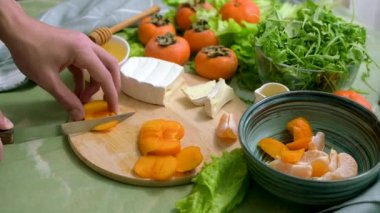 The hands of a young man cut persimmons on a wooden board and a green table to prepare a dietary vegan salad. The process of preparing healthy food. Side view.
