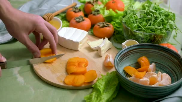 The hands of a young man cut persimmons on a wooden board and a green table to prepare a dietary vegan salad. The process of preparing healthy food. Side view. — ストック動画