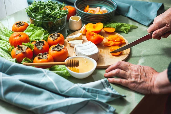 Starsza wegańska kobieta przygotowuje słodką sałatkę z persymony, mandarynki i miękki ser z miodem. Wegańskie zdrowe jedzenie. Dietetyczne odżywianie. Prawdziwe życie. Obraz Stockowy